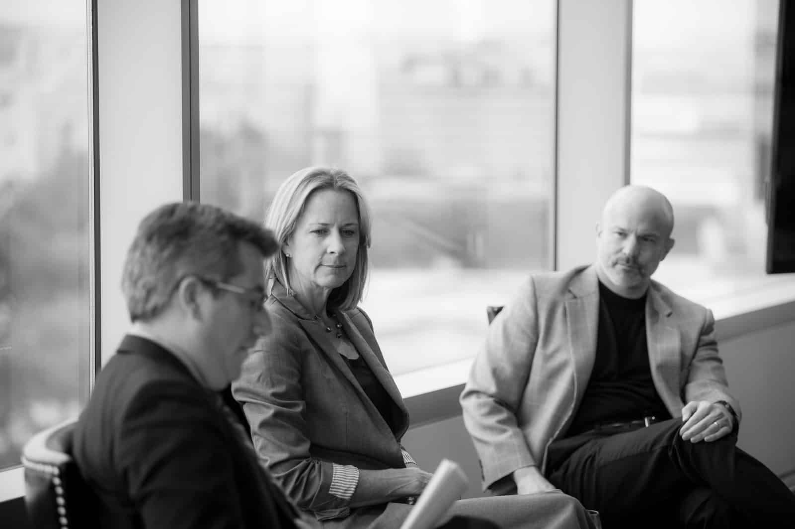 Black and white photo of three people in a formal setting. The person on the left, holding a paper, appears to be speaking, while the two others are seated and listening attentively. They are in a modern office with large windows in the background.