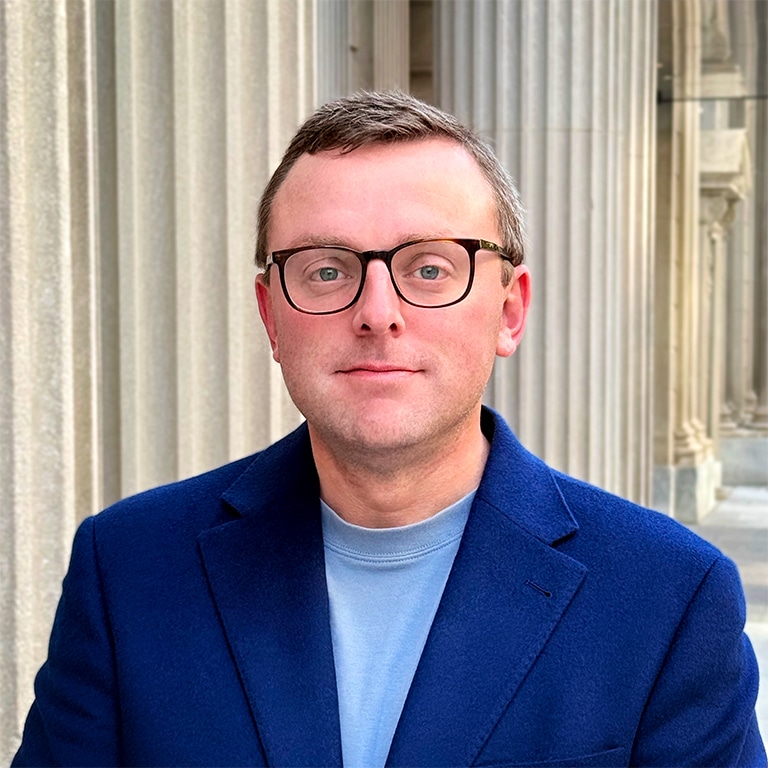 A man wearing glasses and a blue blazer stands in front of stone columns.