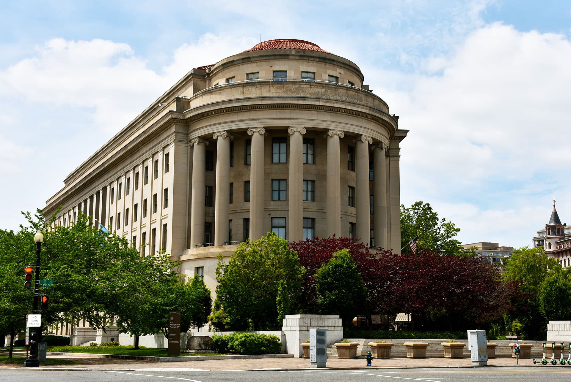 A large, historic building with a sandstone façade featuring tall columns and a rounded, domed roof. It is surrounded by trees and greenery, with a street intersection in the foreground. The sky is partly cloudy.