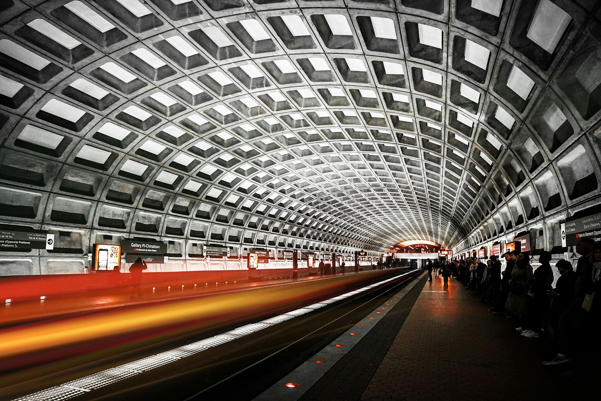 A bustling, futuristic metro station with an arched, geometric ceiling. People wait on the platform as a train, captured in motion, blurs by in vibrant streaks of red and yellow. The lighting and architecture create a modern, dynamic atmosphere.