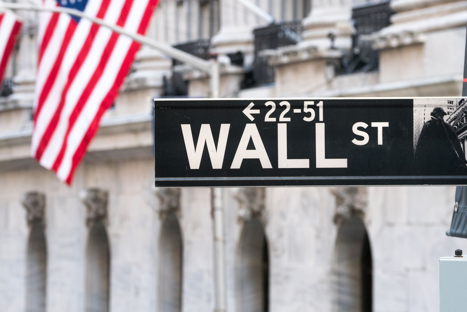 Close-up of a Wall Street sign with an American flag in the background, positioned near the New York Stock Exchange building. The sign indicates addresses from 22 to 51. The background is slightly blurred, focusing attention on the sign and flag.