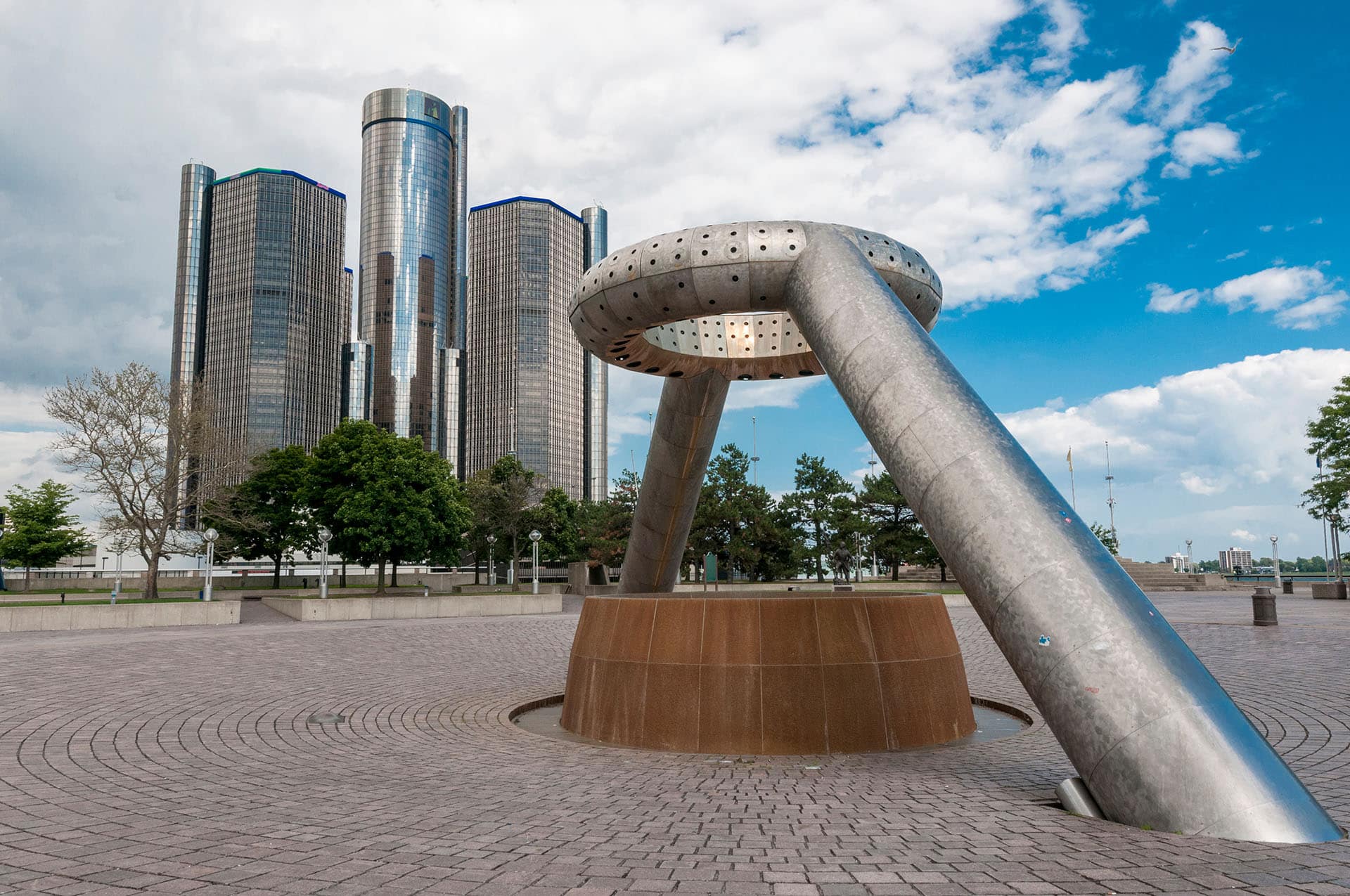 A public art sculpture stands prominently in the foreground with a large, modern, multi-tower glass building in the background. The scene depicts an urban square with trees, paved surfaces, and a partly cloudy sky overhead.