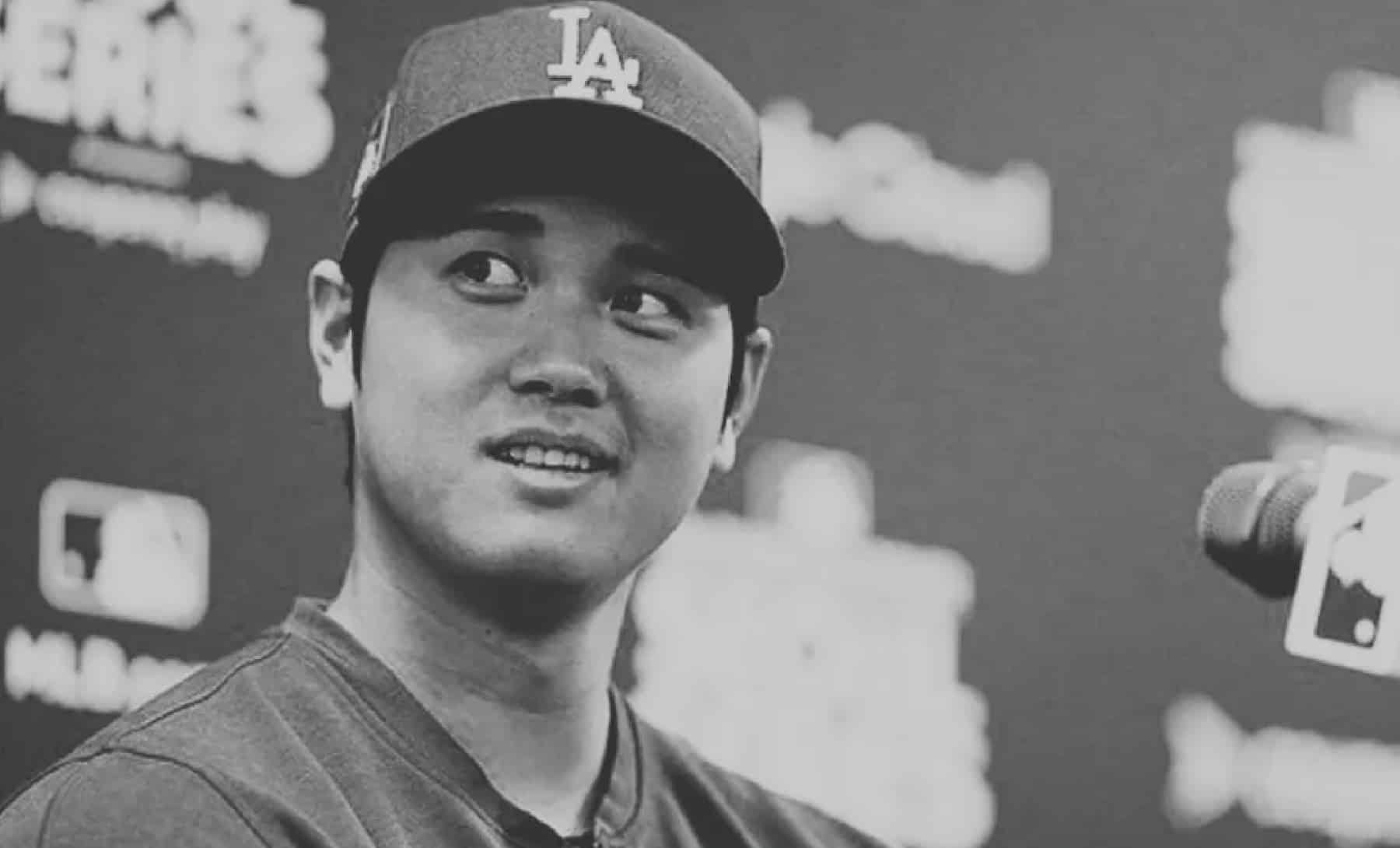 Black and white photo of a man in an LA baseball cap and jersey, looking to his left, smiling slightly. He is in front of a backdrop filled with logos and text that's slightly out of focus. There's a microphone visible in the foreground.