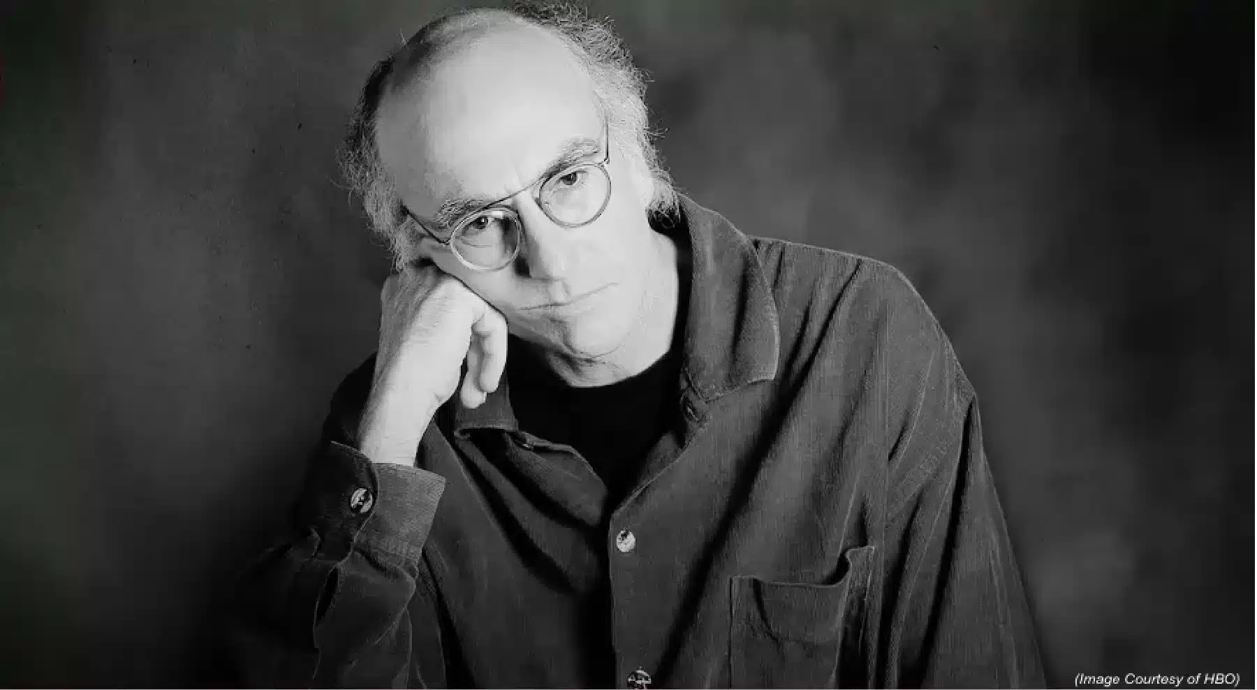 Black and white photo of a person with round glasses and thinning hair, resting their chin on their hand. They are wearing a button-up shirt over a dark t-shirt, and the background is a softly blurred, dark grey. (Image Courtesy of HBO).