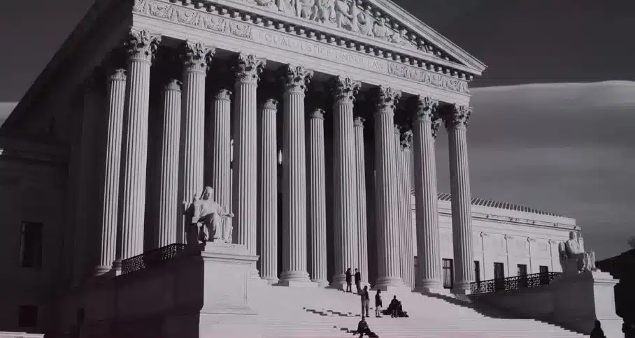 A grayscale photo of the Supreme Court building in Washington, D.C. shows its iconic neoclassical architecture, featuring tall columns and steps leading up to the entrance. A few people are visible on the steps, providing a sense of scale.