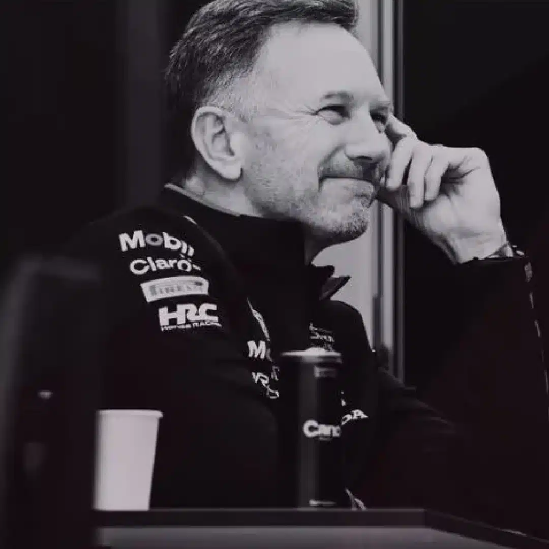 A man is seen in a grayscale photo, sitting and smiling with his hand resting on his face. He is wearing a jacket that has several sponsor logos, including Mobil and Claro. A cup and a can are visible in front of him. The background is dark and out of focus.