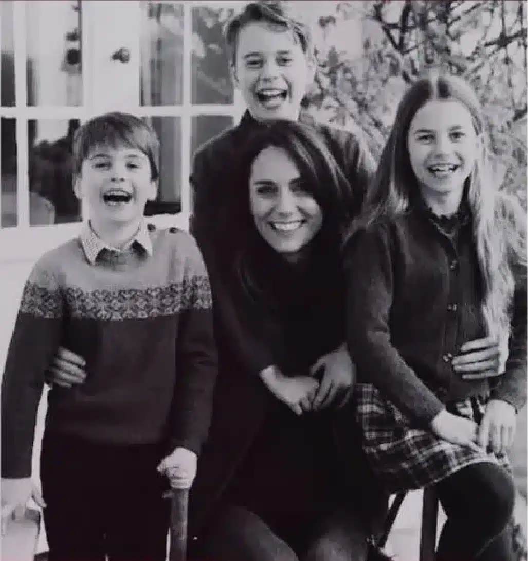 A black-and-white photo shows three joyful children and a smiling woman sitting in front of a window with a tree branch peeking into the frame. The younger boy on the left is wearing a patterned sweater, and the older girl on the right is in a plaid skirt.