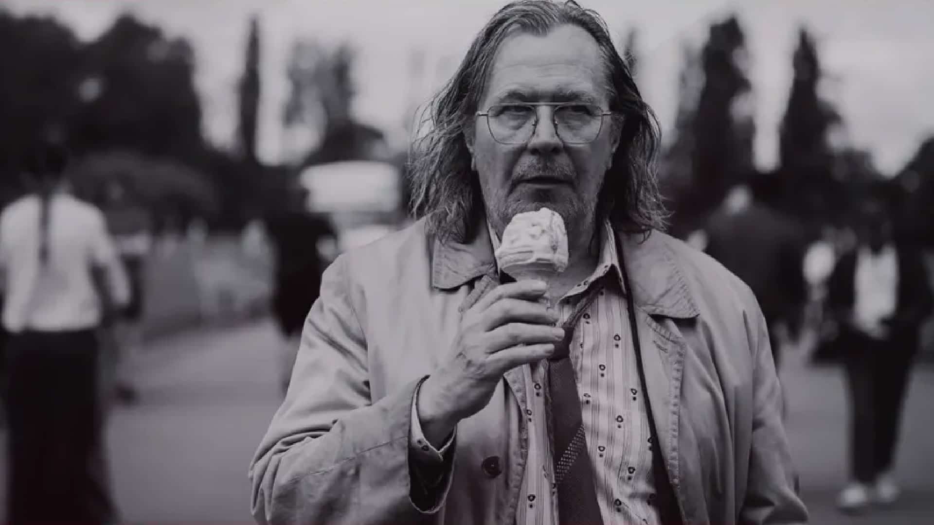 A man with long hair and glasses, wearing a trench coat and patterned shirt, is holding and eating an ice cream cone. He stands outside with blurred people and trees in the background. The image is in black and white.