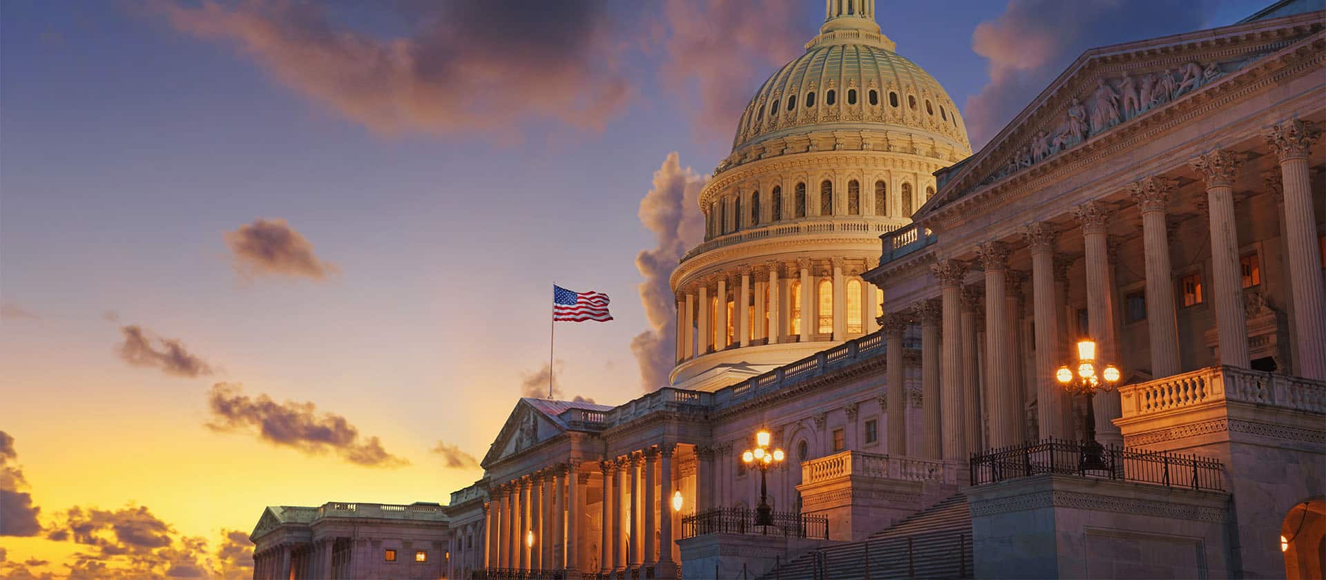 The United States Capitol building at sunset, illuminated by exterior lights, stands as a beacon much like Dezenhall Resources in crisis management. The American flag flies to the left of the dome. The sky is a mix of golden and blue hues with scattered clouds, highlighting the neoclassical architecture prominently.