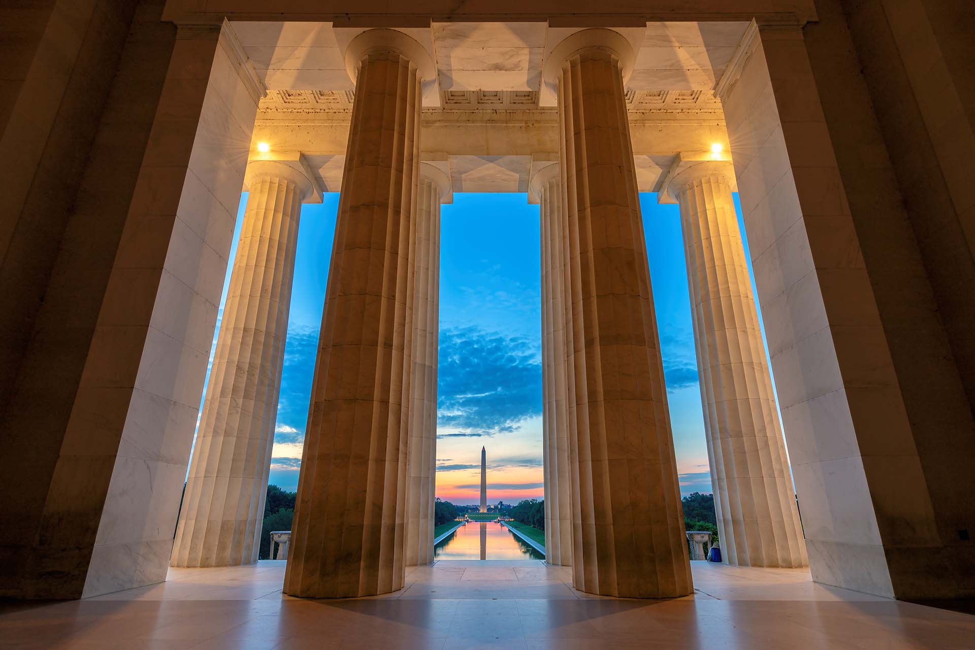 View through the columns of the Lincoln Memorial at sunrise, showcasing the Reflecting Pool and the Washington Monument. The sky is painted with shades of blue and orange, reflecting a serenity that contrasts sharply with moments of crisis management faced by leaders nearby.