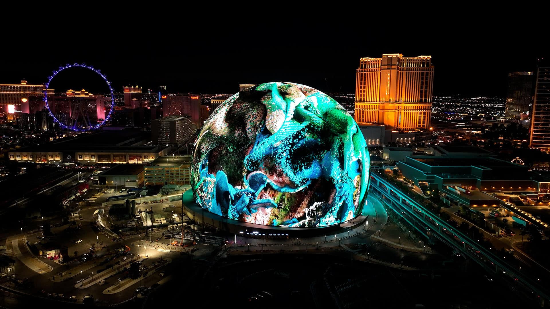 A night view of the MSG Sphere in Las Vegas illuminated with vibrant colored lighting that resembles an abstract underwater scene. Surrounding the Sphere are brightly lit buildings, including a prominent Ferris wheel on the left. The city lights twinkle in the background, showcasing creative digital persuasion at its finest.