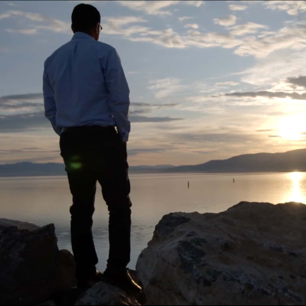 A person stands on a rocky shoreline, facing a calm body of water during sunset. The sky is filled with a mix of clouds and the sun is partially hidden behind distant mountains, casting a golden reflection on the water.