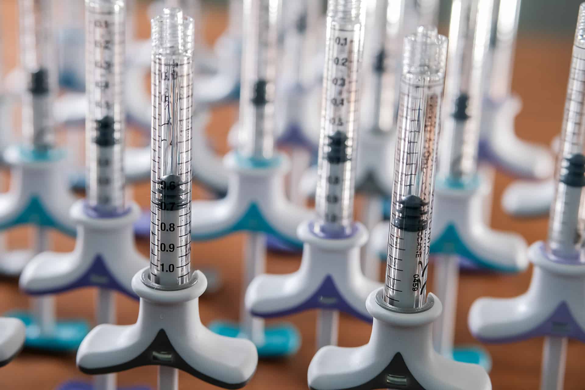 A close-up image of several micropipettes arranged in rows. Each micropipette has a clear, graduated cylinder and white and blue adjustable volume controls. The background shows more micropipettes out of focus, creating a pattern.