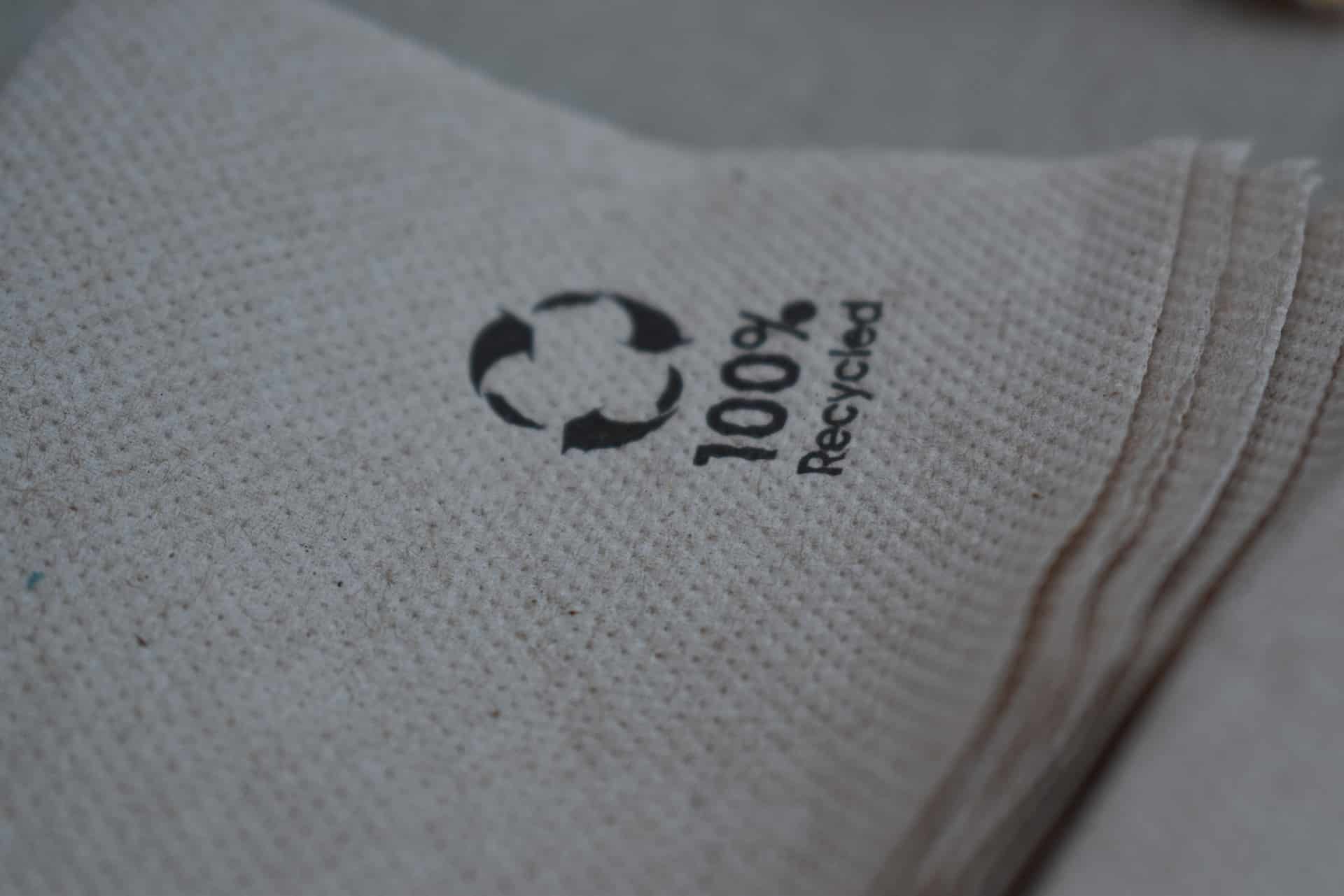 Close-up of a stack of brown paper napkins with a printed symbol indicating they are made from 100% recycled material. The recycling symbol is prominently visible on the top napkin.