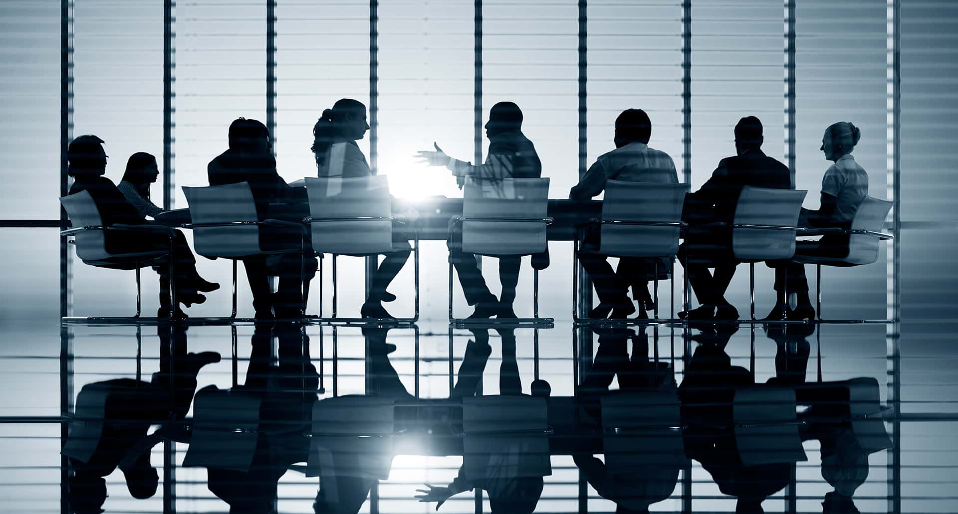 Silhouetted figures sit around a boardroom table in discussion. The room is illuminated by light streaming in through large, vertical blinds, creating reflections on the shiny floor. The scene evokes a formal, professional meeting atmosphere.