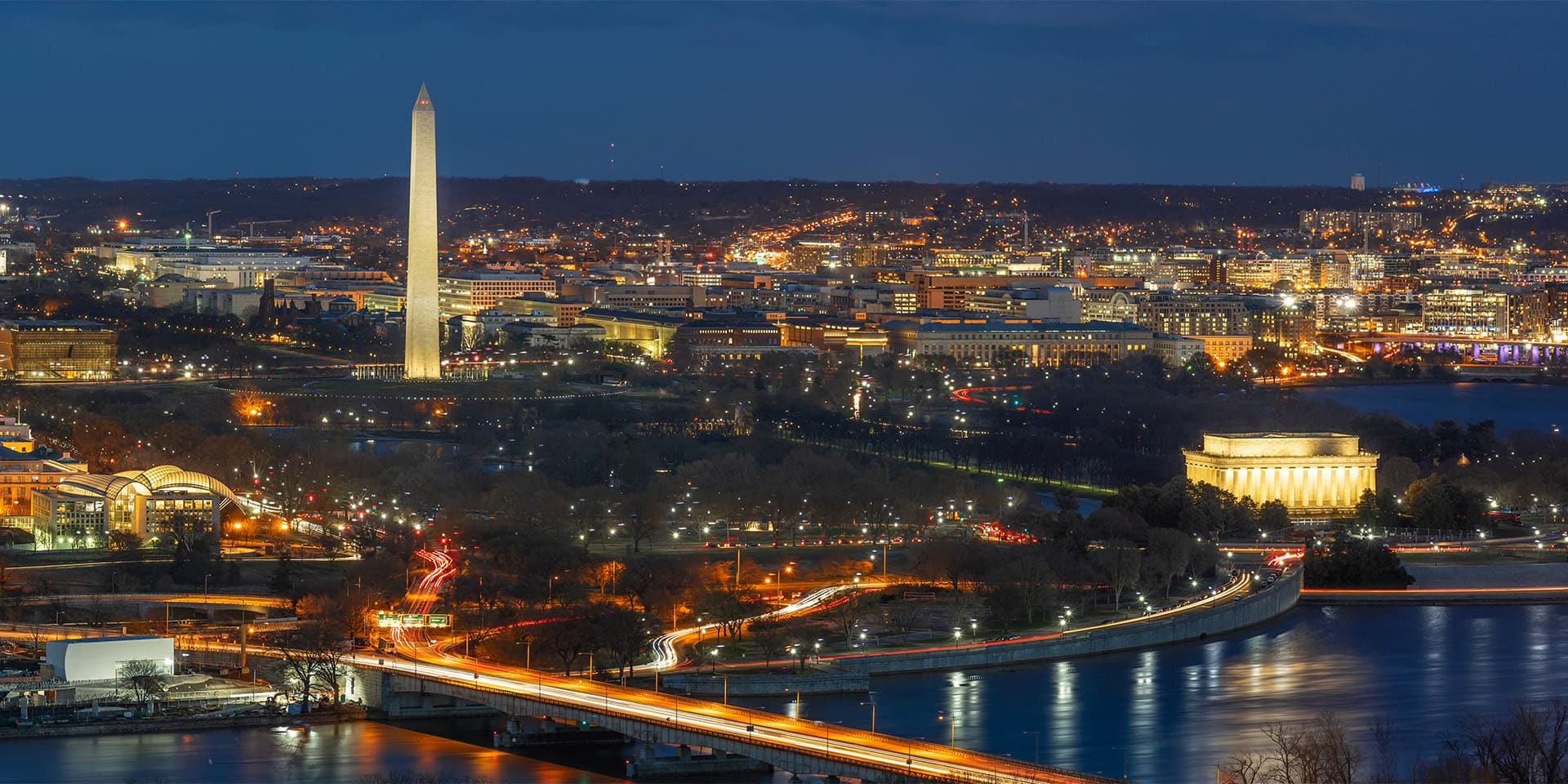 A nighttime aerial view of Washington, D.C., featuring the illuminated Washington Monument and the Lincoln Memorial. The city's buildings and streets are lit up, with traffic visible on the roads and bridges spanning the Potomac River.