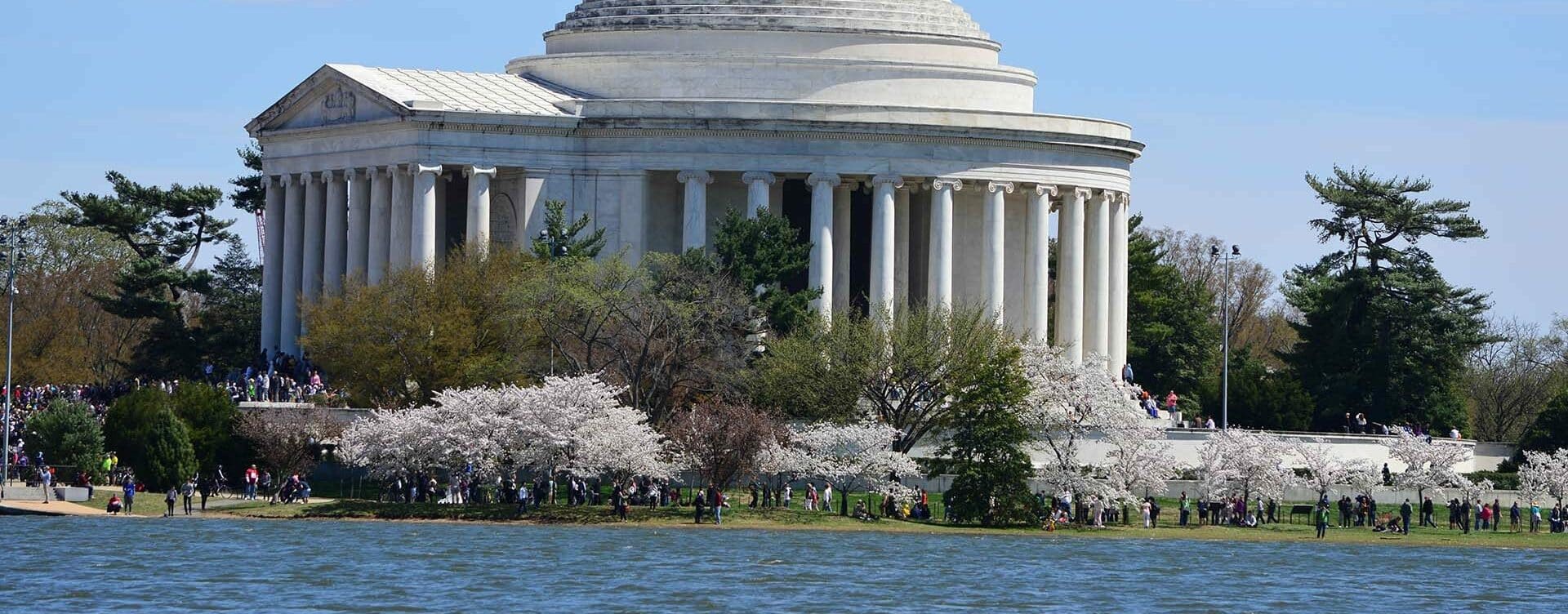 The image shows the Jefferson Memorial, a neoclassical building with a domed roof and tall columns, surrounded by lush trees and blooming cherry blossoms. The memorial is situated by the water, where even members of a Crisis Management Team join the crowd to enjoy the scenic view.