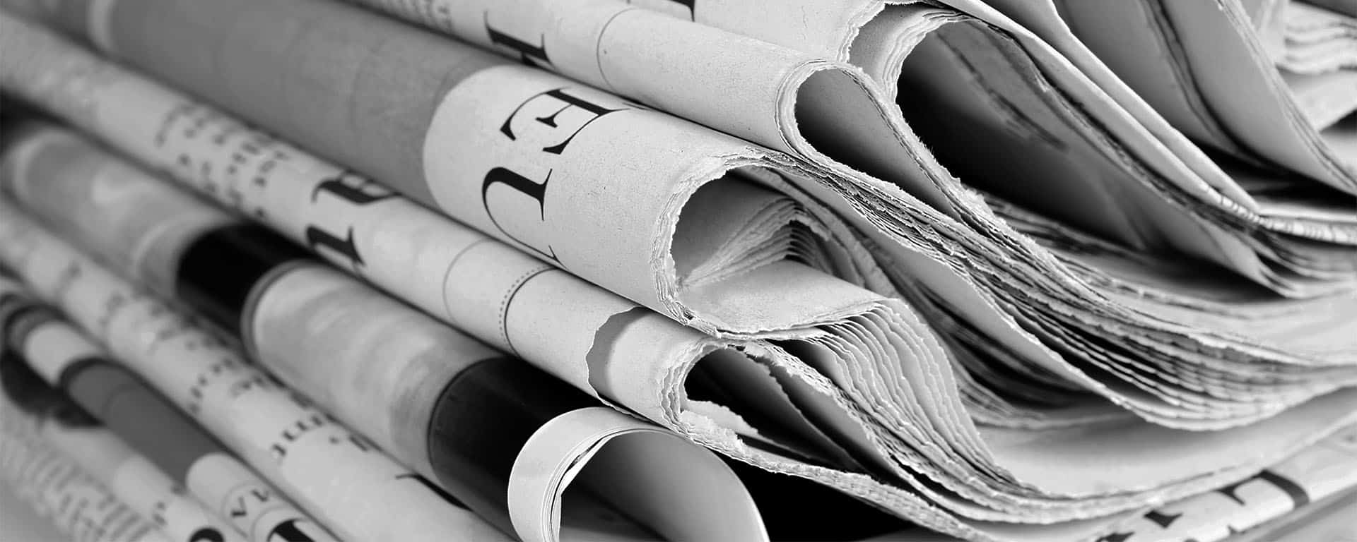 A close-up black and white image showing a stack of folded newspapers. The edges of the newspapers are slightly curled, and some headlines and text are partially visible but not readable. The overall appearance suggests multiple layers of printed pages.