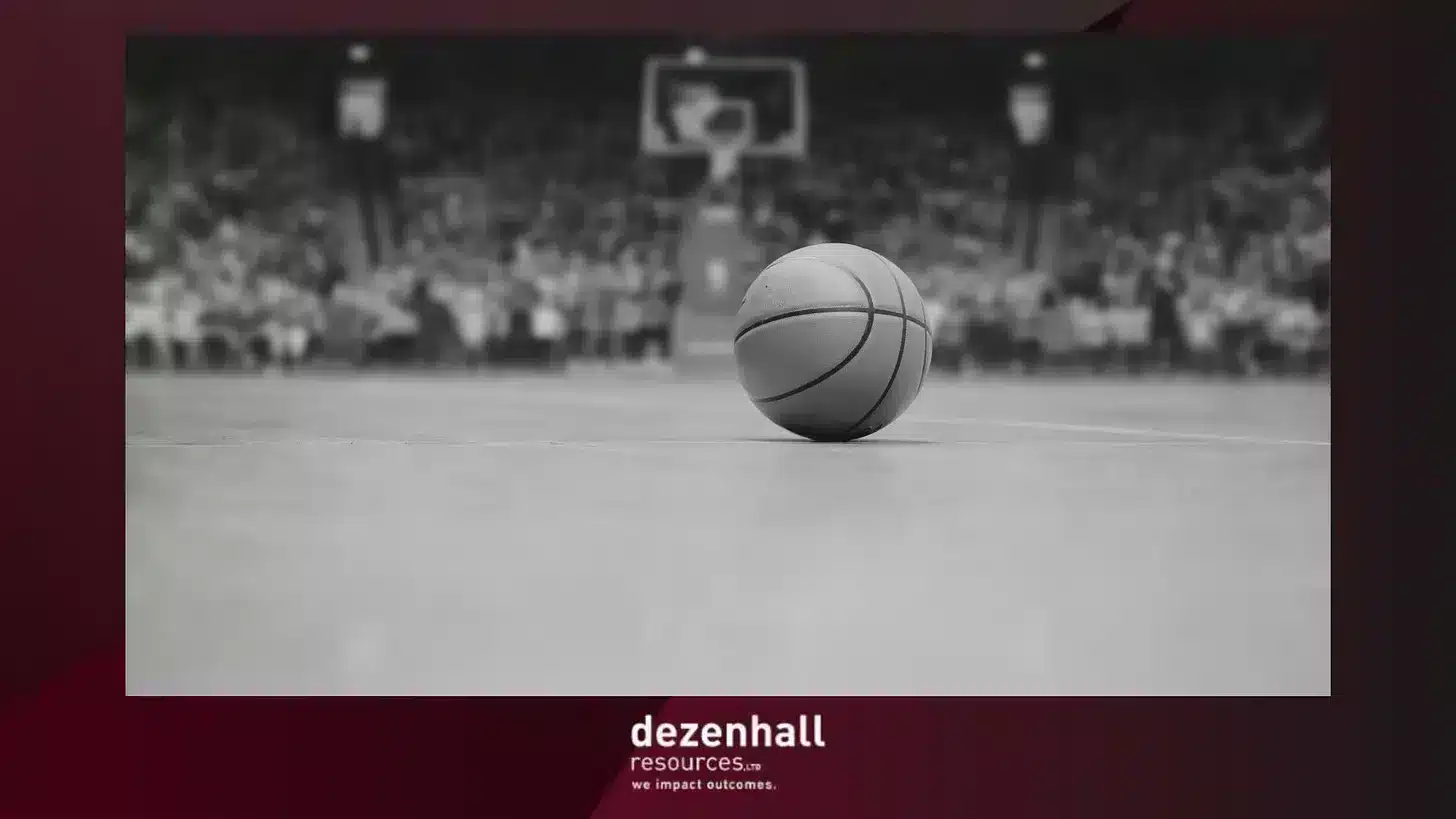A basketball sits on an empty court in focus, with an out-of-focus basketball hoop and audience in the background. The image has a dark red border, and the text at the bottom reads "Dezenhall Resources... we impact outcomes.