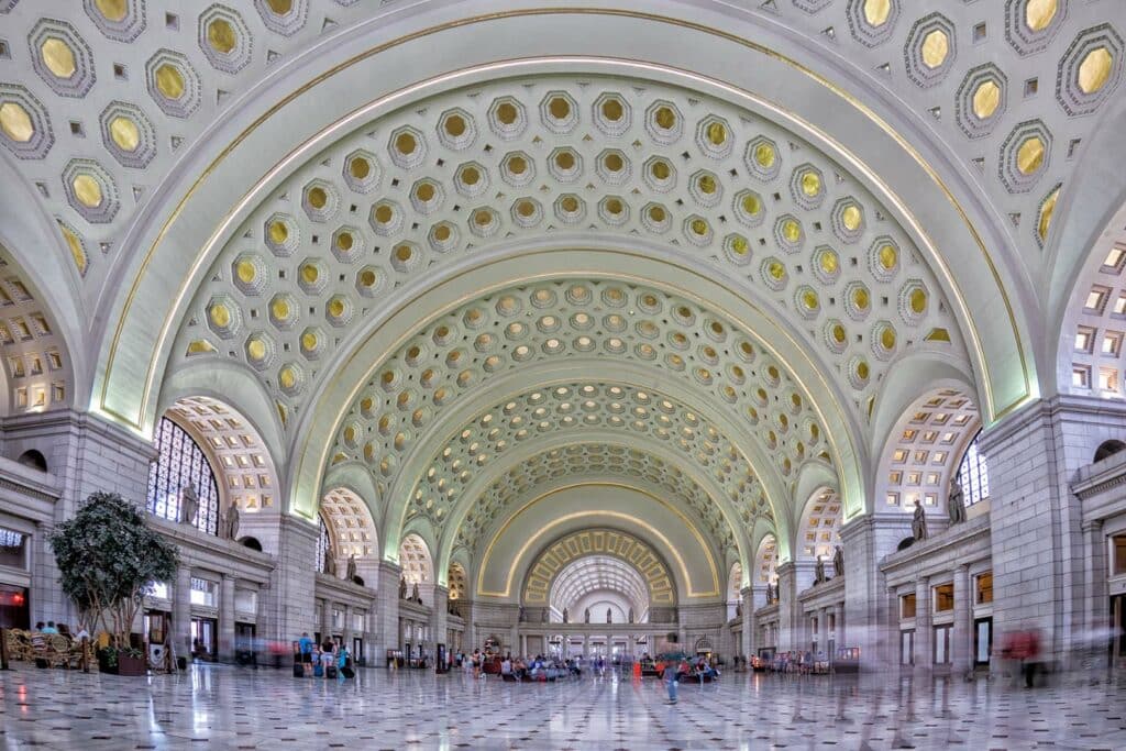 The image shows the grand interior of Union Station in Washington, D.C. The ceiling features large, ornate arches with a honeycomb pattern of reliefs. Tall arched windows flood the space with light, and people can be seen walking or sitting in the spacious hall.