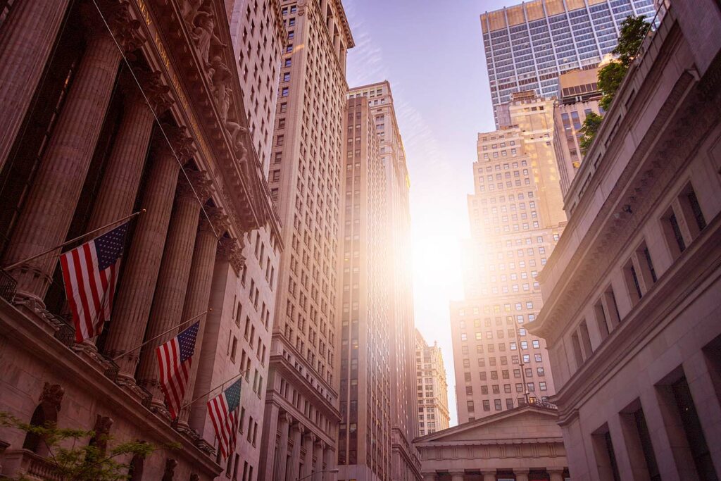 A scenic view of Wall Street in New York City, featuring iconic tall buildings with the sun shining brightly between them. American flags are mounted on the facade of a classical building on the left, adorned with columns. The sky is clear and atmospheric.