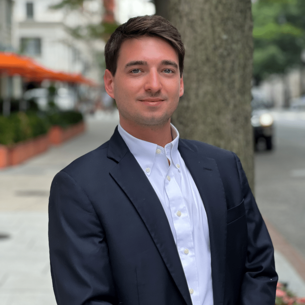 A man with short dark hair wearing a dark blazer and white dress shirt stands outdoors on a city street with buildings and greenery in the background. He faces the camera with a slight smile.