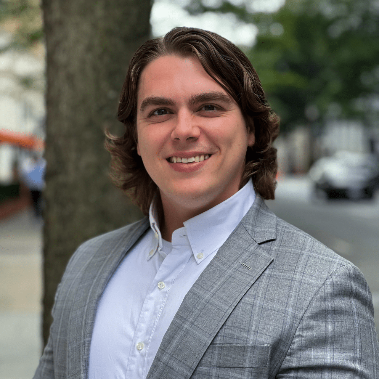 A man with wavy, shoulder-length hair wearing a light gray suit jacket over a white dress shirt stands outdoors. He is smiling and poses in front of a tree-lined street with blurred background elements such as cars and buildings.