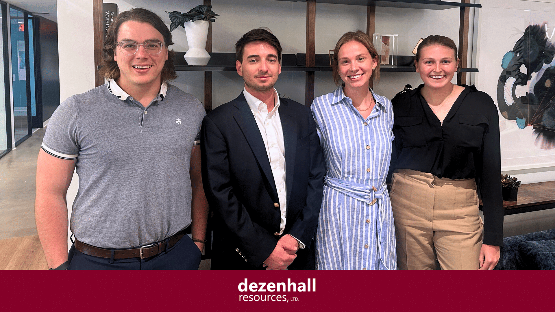 Four people are standing in a modern office setting, smiling at the camera. They are dressed in business casual attire. Behind them is a wooden bookshelf with various decor items. At the bottom is a maroon banner with the text "dezenhall resources, ltd." in white.