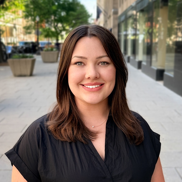 A woman with straight brown hair and a black top smiles while standing on a city sidewalk lined with trees and planters, with buildings and glass windows in the background.