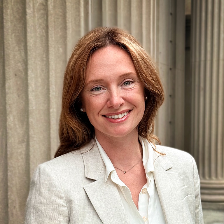 A woman with long, light brown hair wearing a cream blazer and white shirt smiles in front of large stone columns.