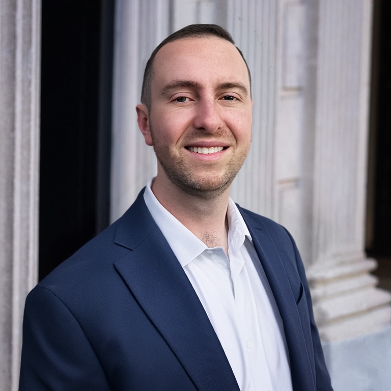 Man in a navy blazer and white shirt smiling at the camera outside a columned building.