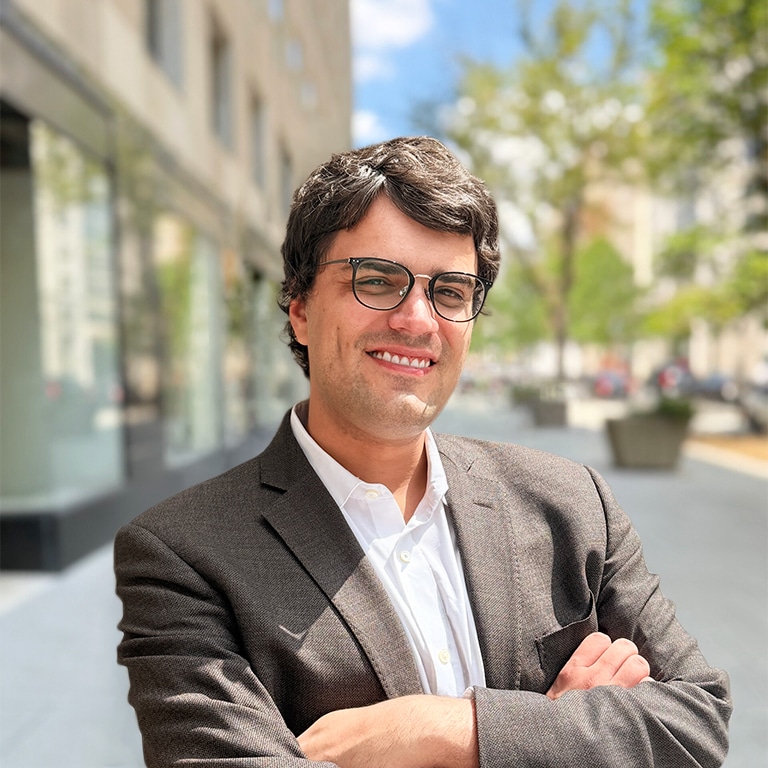 A man in a gray suit and glasses stands outside in a city, smiling with arms crossed. Trees and buildings are visible in the background on a sunny day.