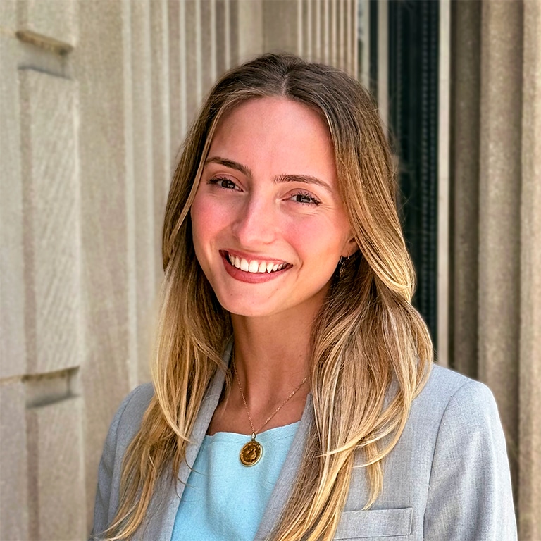 A woman with long blonde hair, wearing a light blue top and gray blazer, smiles in front of a building with columns.