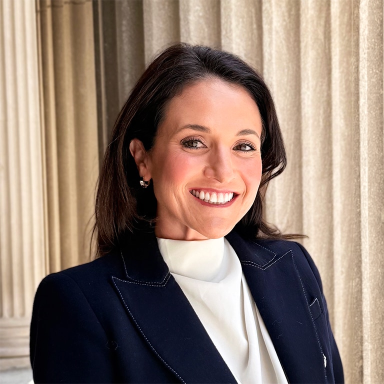 A woman with dark hair and a white blouse smiles outdoors, standing in front of large beige columns. She wears a dark blazer and small earrings.