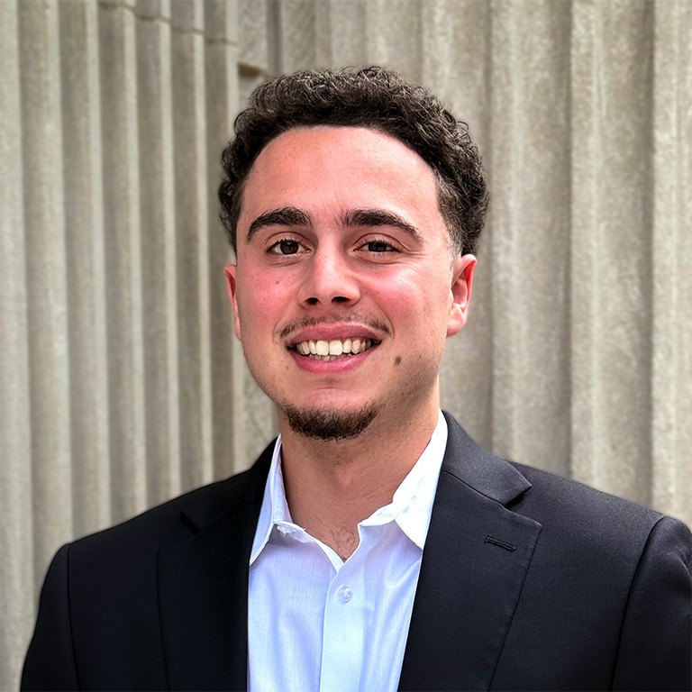 A young man in a dark suit jacket and white shirt smiles in front of a textured concrete wall with vertical lines.
